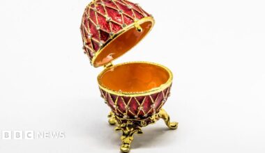 Open red and gold Fabergé-style egg with diamond-patterned enamel and crystal detailing, displayed on ornate gold legs against a white background.