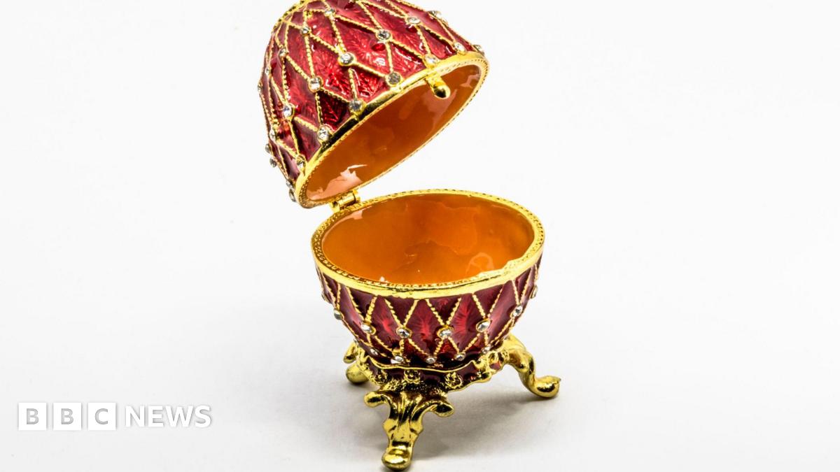 Open red and gold Fabergé-style egg with diamond-patterned enamel and crystal detailing, displayed on ornate gold legs against a white background.