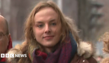 Sandy Peggie has shoulder-length blonde hair and is wearing a blue jacket over a white top