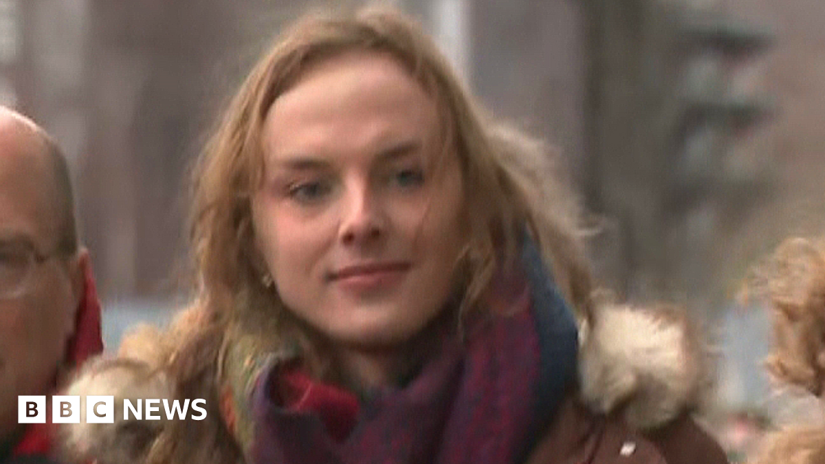 Sandy Peggie has shoulder-length blonde hair and is wearing a blue jacket over a white top