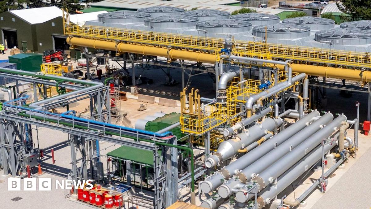 The United Downs power plant in Cornwall is shown, there are various silver pipes surrounding a central plant that has a series of large fan type structures surrounded by yellow pipes. There are trees in the background and the sky is blue.