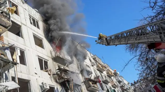 Smoke rises from a building while a firefighter on a crane sprays water from a hose