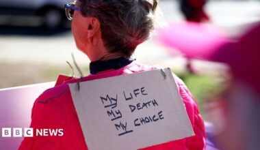 A person stands with their back to the camera and a sign hanging over her neck reading 'my life, my death, my choice, with bokeh background and foreground detail, outside parliament in London in September.