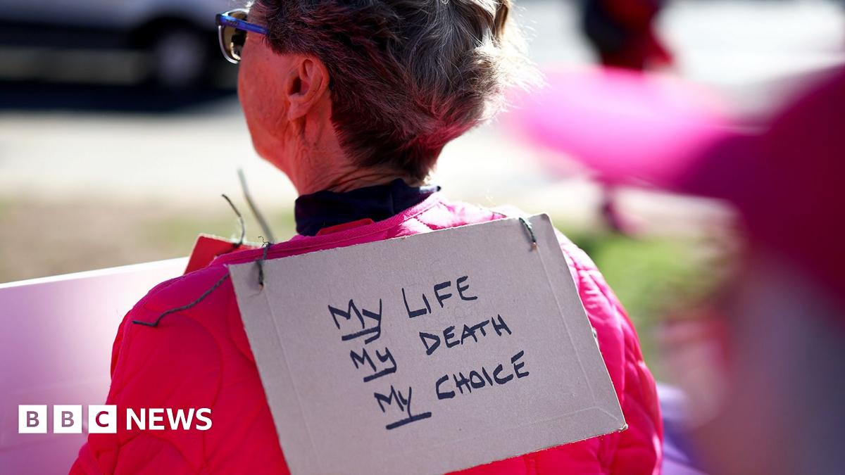 A person stands with their back to the camera and a sign hanging over her neck reading 'my life, my death, my choice, with bokeh background and foreground detail, outside parliament in London in September.