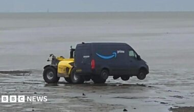 A white van is positioned heading downhill off a city street. It has mounted the pavement and appears to be stuck. Two pedestrians are walking round the back of the van.