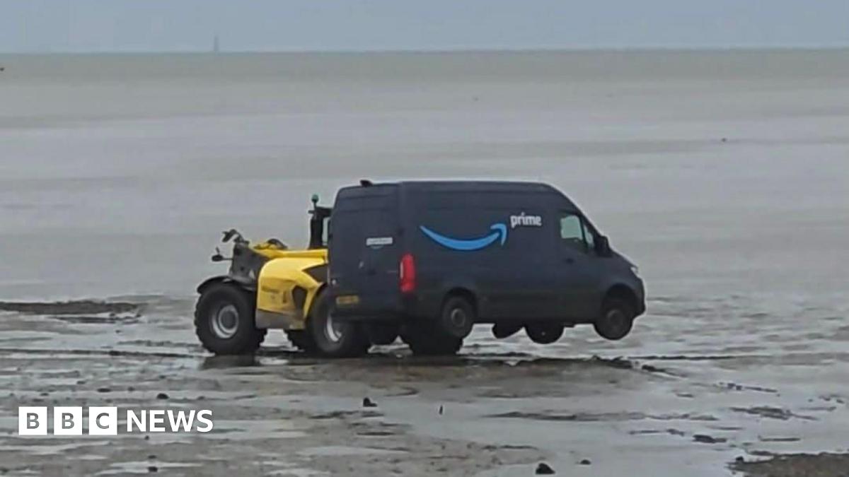A white van is positioned heading downhill off a city street. It has mounted the pavement and appears to be stuck. Two pedestrians are walking round the back of the van.