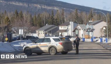 Policeman stands beside police car parked half across a residential road with mountain behind in Tumbler Ridge, British Columbia
