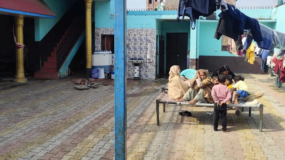 A quiet courtyard inside a village home in Umri. Several women and children sit together on a woven cot while one child stands nearby. Clothes hang on lines overhead, and the surrounding walls are painted blue and turquoise, with doorways and a staircase leading into the house