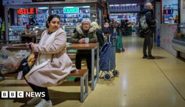 People do their shopping in Gorton Market on January 28, 2026 in Gorton, United Kingdom. A woman can be seen sat on a bench with her shopping while an older woman sits with a cup of tea behind her. Others can be seen looking at stalls.