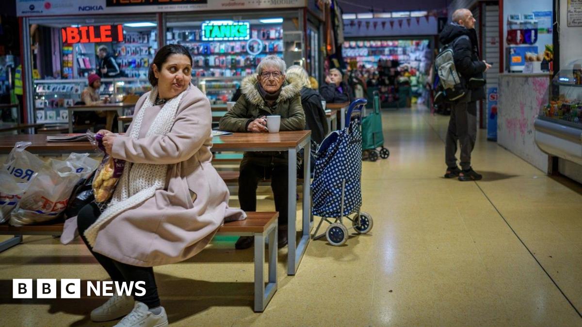 People do their shopping in Gorton Market on January 28, 2026 in Gorton, United Kingdom. A woman can be seen sat on a bench with her shopping while an older woman sits with a cup of tea behind her. Others can be seen looking at stalls.