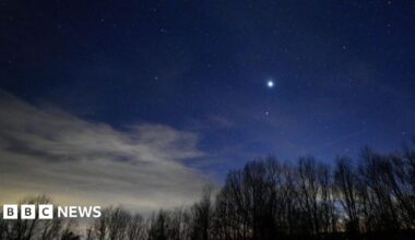 A father and two children sit on a wooden deck at dusk, facing a deep blue evening sky. All three are pointing upward at faint stars, and a small telescope on a tripod stands beside them as they stargaze together.