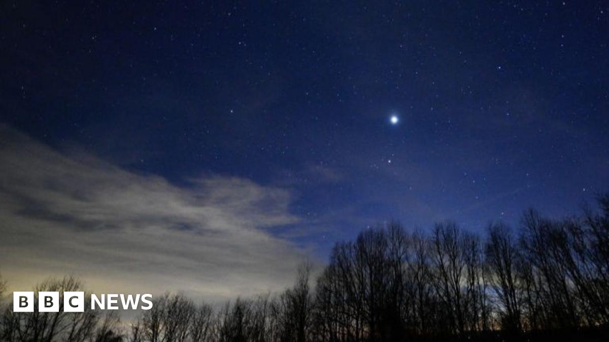 A father and two children sit on a wooden deck at dusk, facing a deep blue evening sky. All three are pointing upward at faint stars, and a small telescope on a tripod stands beside them as they stargaze together.