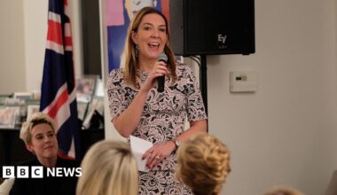 Dame Antonia Romeo has long brown hair and is wearing a floral patterned dress in pale pink and blue. She is speaking into a microphone