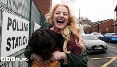 Hannah Spencer stands in front of a polling station sign attached to a metal fence. Spencer is smiling, holding an orange coloured bag and wearing a multi-coloured coat.