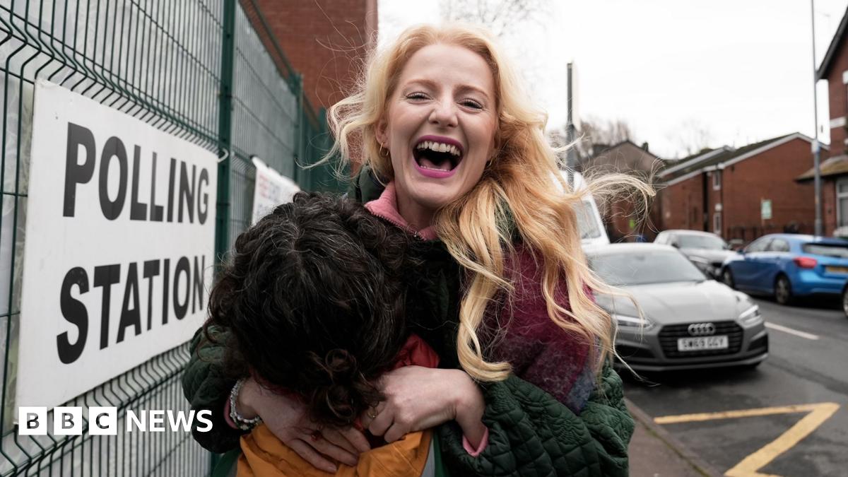 Hannah Spencer stands in front of a polling station sign attached to a metal fence. Spencer is smiling, holding an orange coloured bag and wearing a multi-coloured coat.