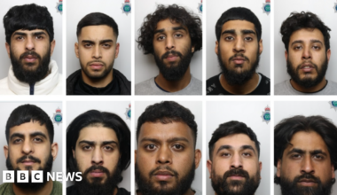 Ten men with dark hair and beards in front of a grey background