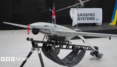 A Ukrspecsystems drone with a shark face and design on its nose propped up on a black metal stand. In the background there is a white banner which reads Ukrspecsystems and a Ukraine flag.