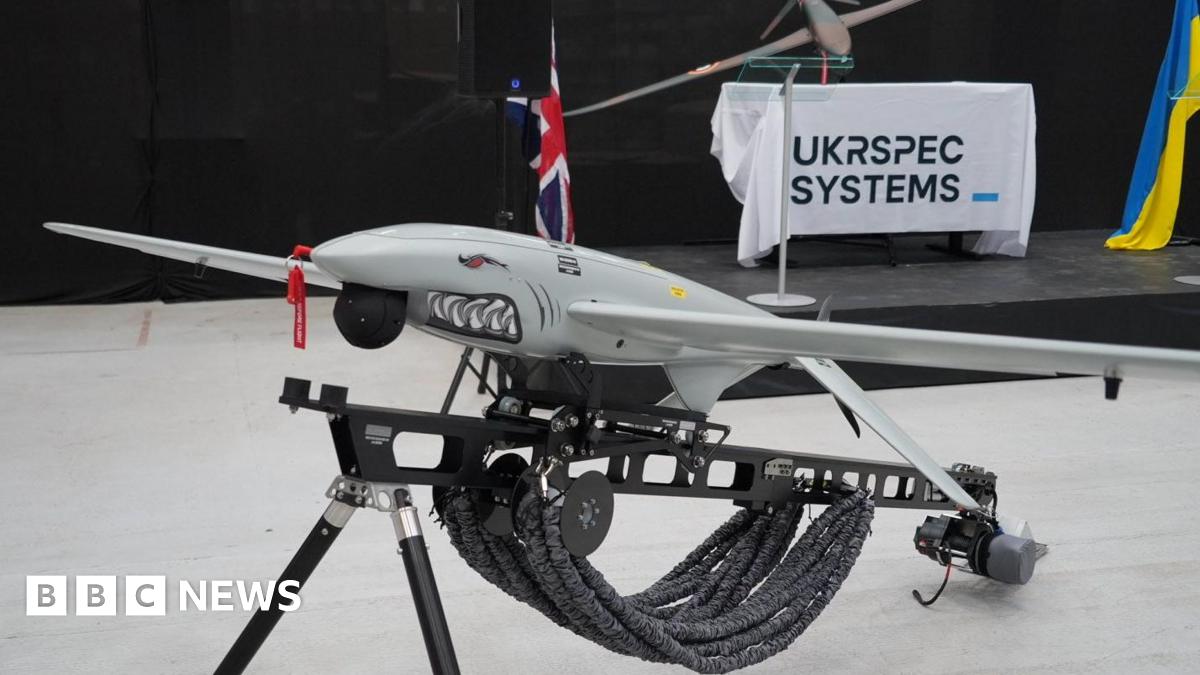 A Ukrspecsystems drone with a shark face and design on its nose propped up on a black metal stand. In the background there is a white banner which reads Ukrspecsystems and a Ukraine flag.