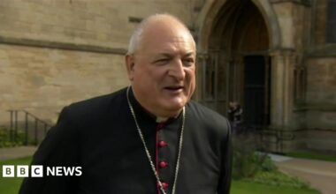 The photo shows a man looking to the right-hand side of the camera. He is wearing a black cassock and a silver chain around his neck. He's standing in front of a church.