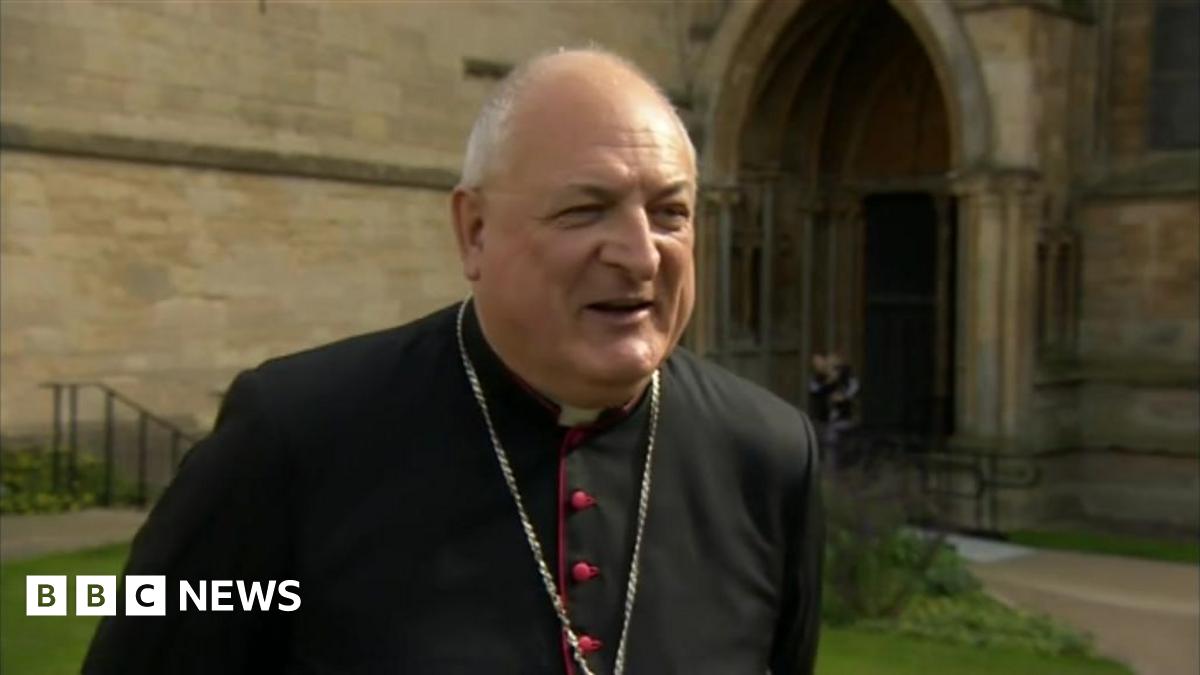 The photo shows a man looking to the right-hand side of the camera. He is wearing a black cassock and a silver chain around his neck. He's standing in front of a church.