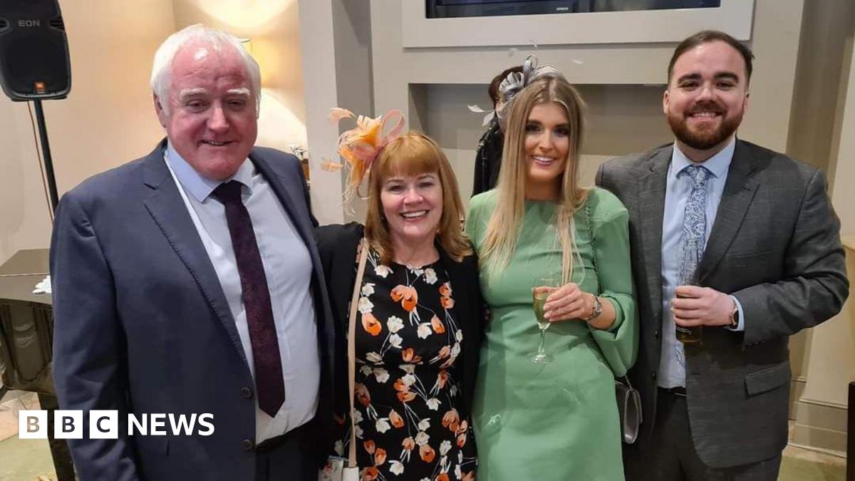 Four people standing next to each other in a photo. They are wearing smart clothes and smiling at the camera. Two men are wearing jackets and ties; the two women are wearing dresses and fascinators.