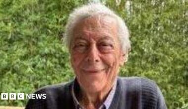 An elderly man with grey hair and wearing a dark blue jumper smiles at the camera.