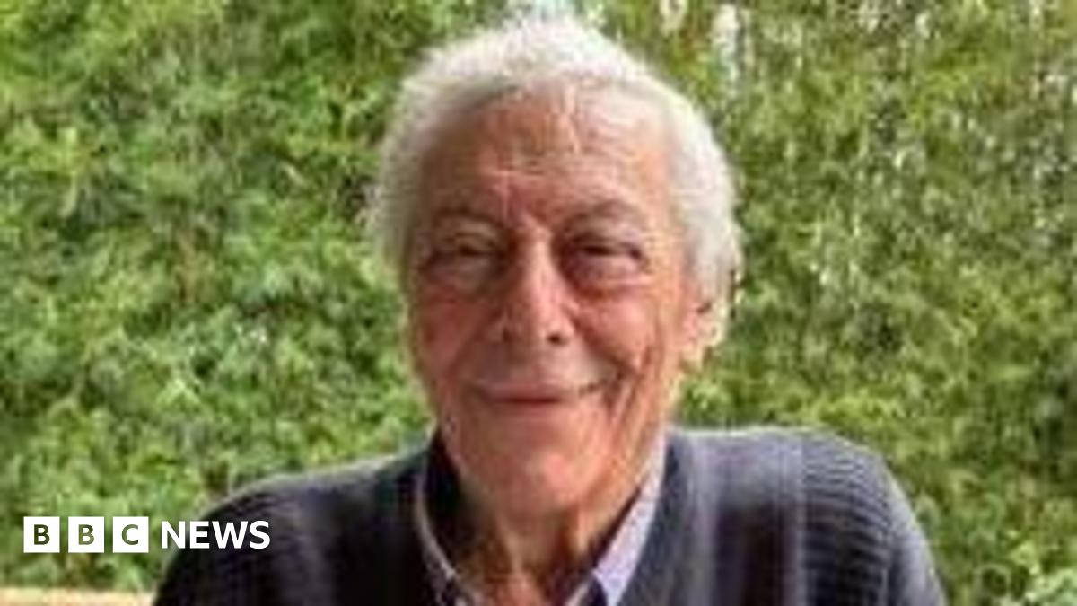 An elderly man with grey hair and wearing a dark blue jumper smiles at the camera.