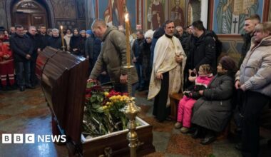 A man pays respects to a recent casualty, with the open coffin with flowers in the middle of a church hall, and other mourners all around