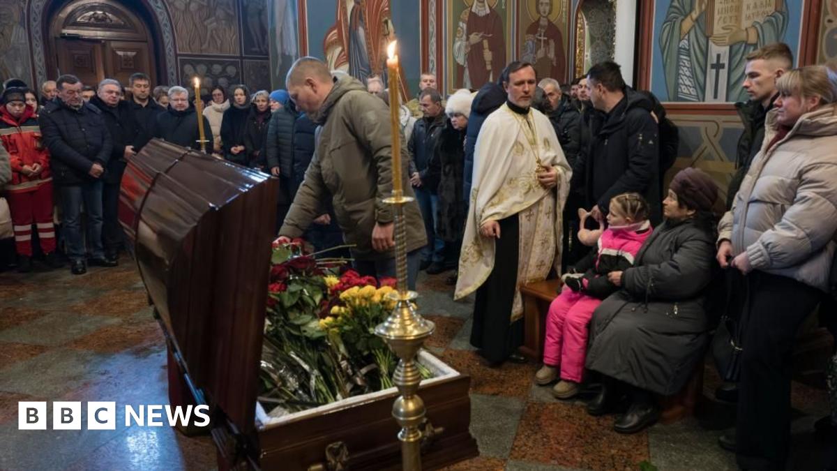 A man pays respects to a recent casualty, with the open coffin with flowers in the middle of a church hall, and other mourners all around