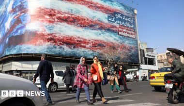 Iranians walk past a billboard warning the US not to attack Iran at a square in central Tehran, Iran (25 February 2026)