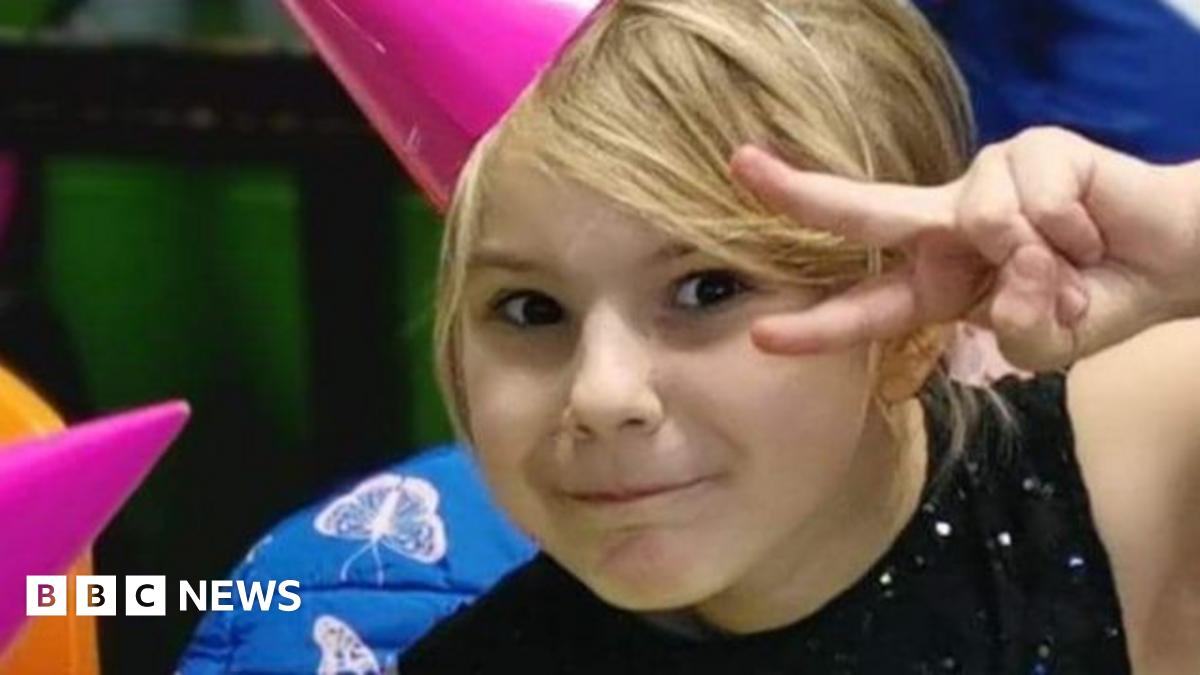 A girl with blonde hair with a side fringe is pulling a cheeky expression at the camera and pulling a peace sign. She is wearing a pink party hat and a black sparkly top.