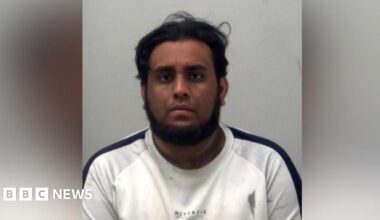 A man wearing a white top with a black strip down the shoulder. He has a beard and he is looking at the camera. It is a custody photo.