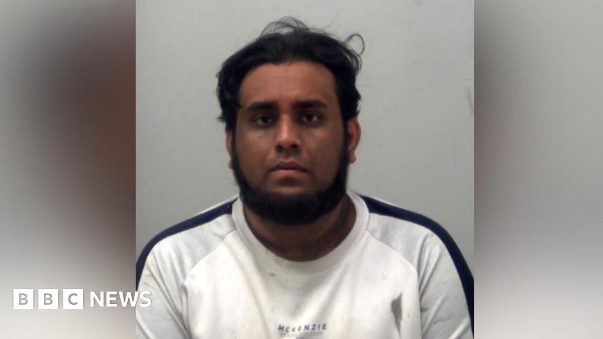 A man wearing a white top with a black strip down the shoulder. He has a beard and he is looking at the camera. It is a custody photo.