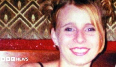 A grainy image of Victoria Hall smiling at the camera, with her blonde hair swept down to one side. She is wearing bright lipstick and an earring.