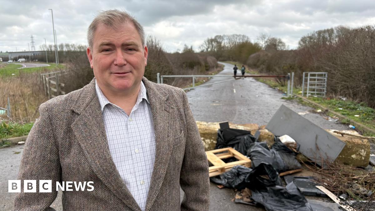 Philip Spicer with short brown hair, wearing a brown jacket over a white shirt. He is standing beside some rubbish dumped in front of a gated road.  It includes black-bagged waste, pieces of wood and tree branches