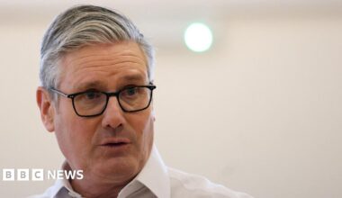 A close-up shot of Keir Starmer, wearing black glasses and a white shirt