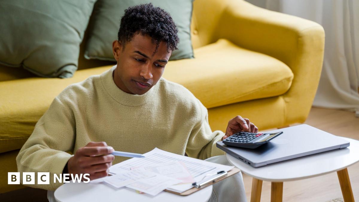 Stock photo shows a person looking down at a clipboad with bills and receipts while holding a pen and using a calculator in their living room at home.