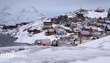Houses of the old town are pictured in Nuuk, western Greenland. Around them are snowy rolling hills and a small mountain in the distance. The houses have snowy roofs, and ar different colours -- red, yellow, teal and blue, and are densly packed by the shore.