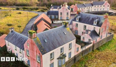 Close up drone shot of a street in the village. The houses are white with black bricked roofs. They are derelict with red marks on their sides.