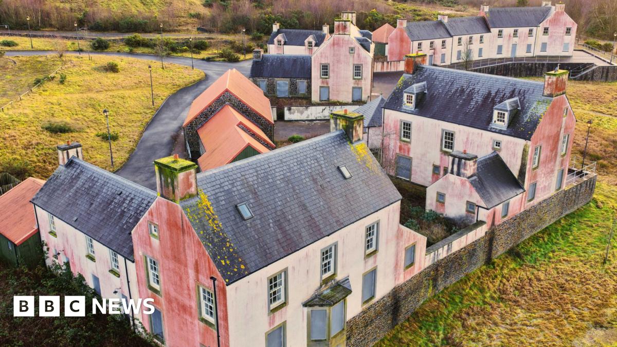 Close up drone shot of a street in the village. The houses are white with black bricked roofs. They are derelict with red marks on their sides.