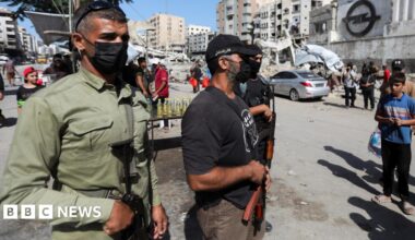 Three Hamas militants wearing face masks and carrying automatic weapons stand guard on a destroyed street in downtown Gaza.