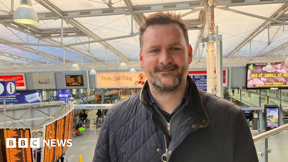 Photograph of Simon Bennett, the station manager at Manchester Piccadilly Station. The image shows the departure board and the platforms in the background.