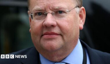 A man in a blue shirt and tie, with a dark navy blazer. He wears glasses and has grey hair.