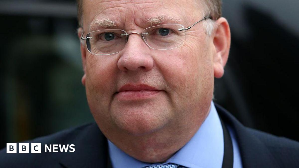 A man in a blue shirt and tie, with a dark navy blazer. He wears glasses and has grey hair.