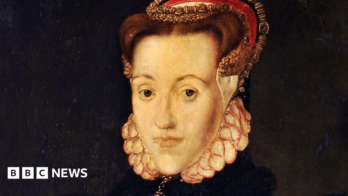 A woman with light-grey hair in a bob wears a blue jacket and stands on a promenade staring across the grey-brown waters of a wide river estuary. White railings and a large glass building can be seen to her right. The sky is blue and dotted with white clouds.