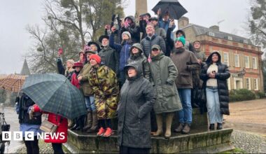 King Charles points at the town crier, who is wearing a red coat and holding the Union flag.