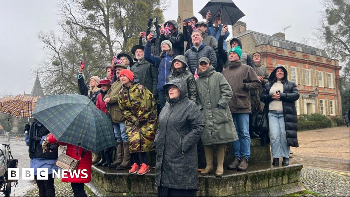 King Charles points at the town crier, who is wearing a red coat and holding the Union flag.