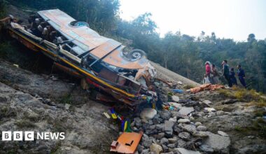 A wide shot of the bus which is lying on its side on the river bed, with the wheels facing up at a slant. To the right of it, are five people looking up. There are trees in the background. It is day