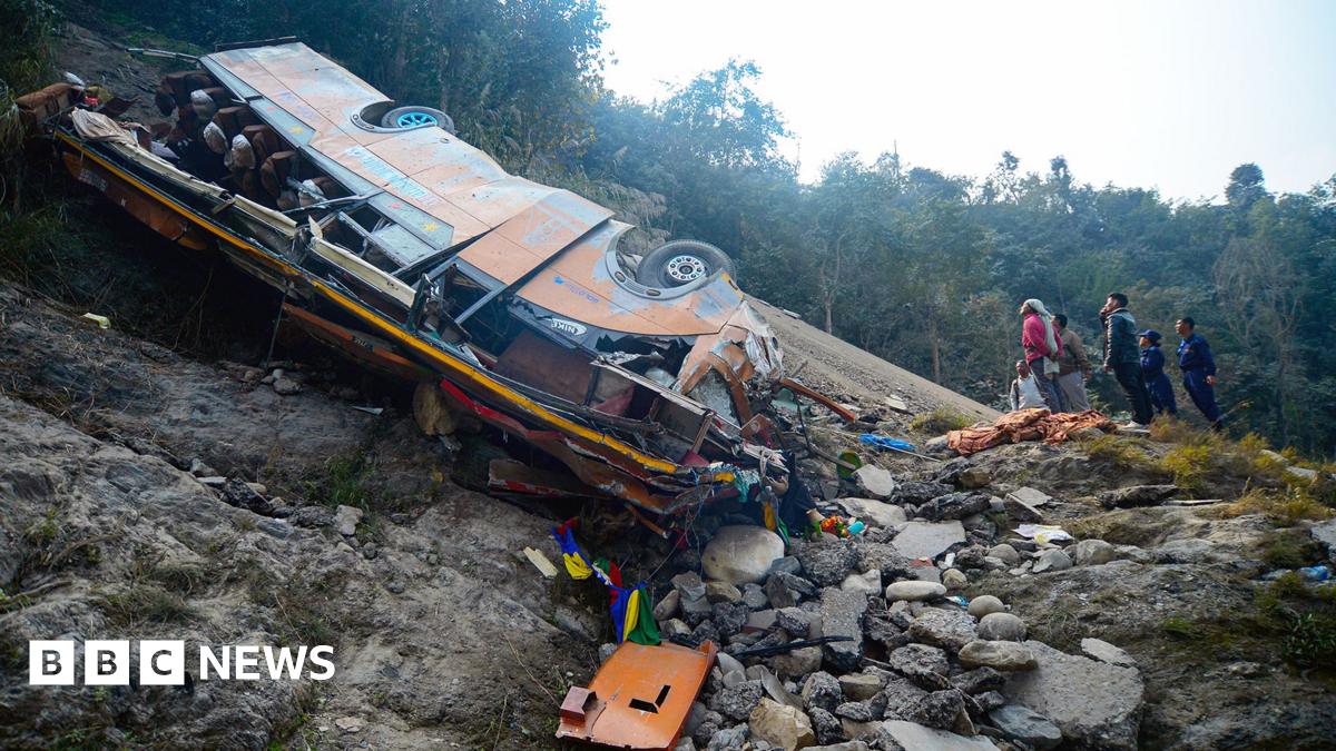 A wide shot of the bus which is lying on its side on the river bed, with the wheels facing up at a slant. To the right of it, are five people looking up. There are trees in the background. It is day