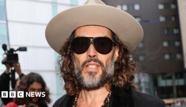 Russell Brand arriving at Southwark Crown Court wearing a light grey fedora hat, dark glasses, a leopard print shirt unbuttoned halfway down his chest, a cross on a necklace, and a dark jacket